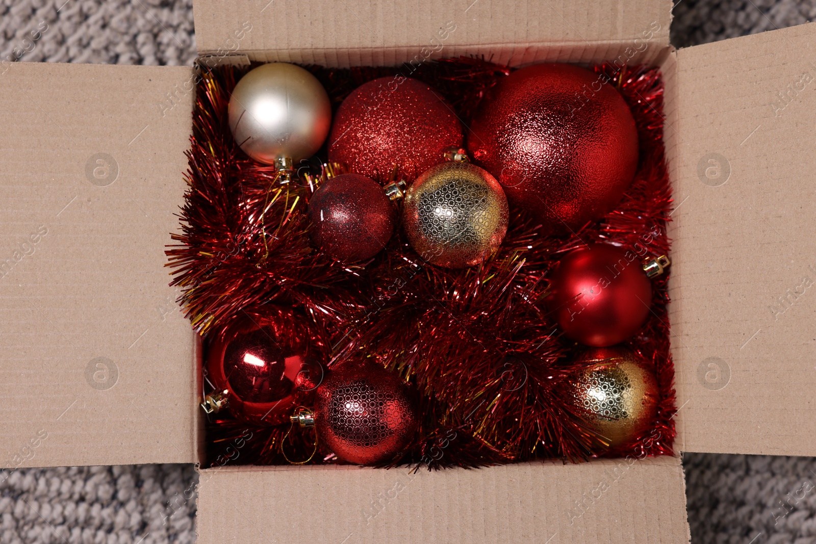 Photo of Shiny tinsels and baubles in cardboard box on carpet, top view