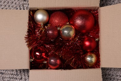 Photo of Shiny tinsels and baubles in cardboard box on carpet, top view