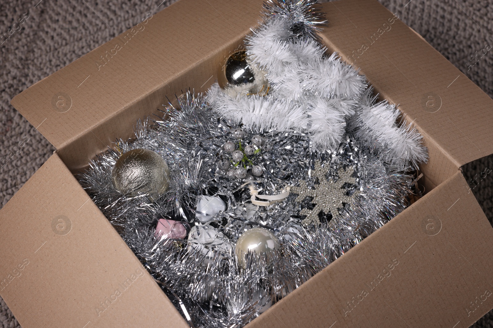 Photo of Shiny tinsels, baubles and other Christmas decor in cardboard box on carpet, closeup