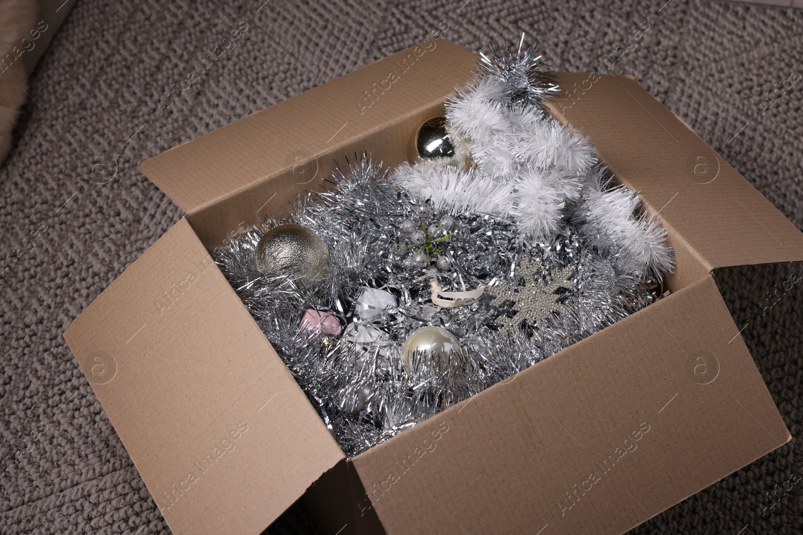 Photo of Shiny tinsels, baubles and other Christmas decor in cardboard box on carpet, closeup