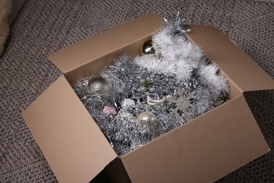 Photo of Shiny tinsels, baubles and other Christmas decor in cardboard box on carpet, closeup