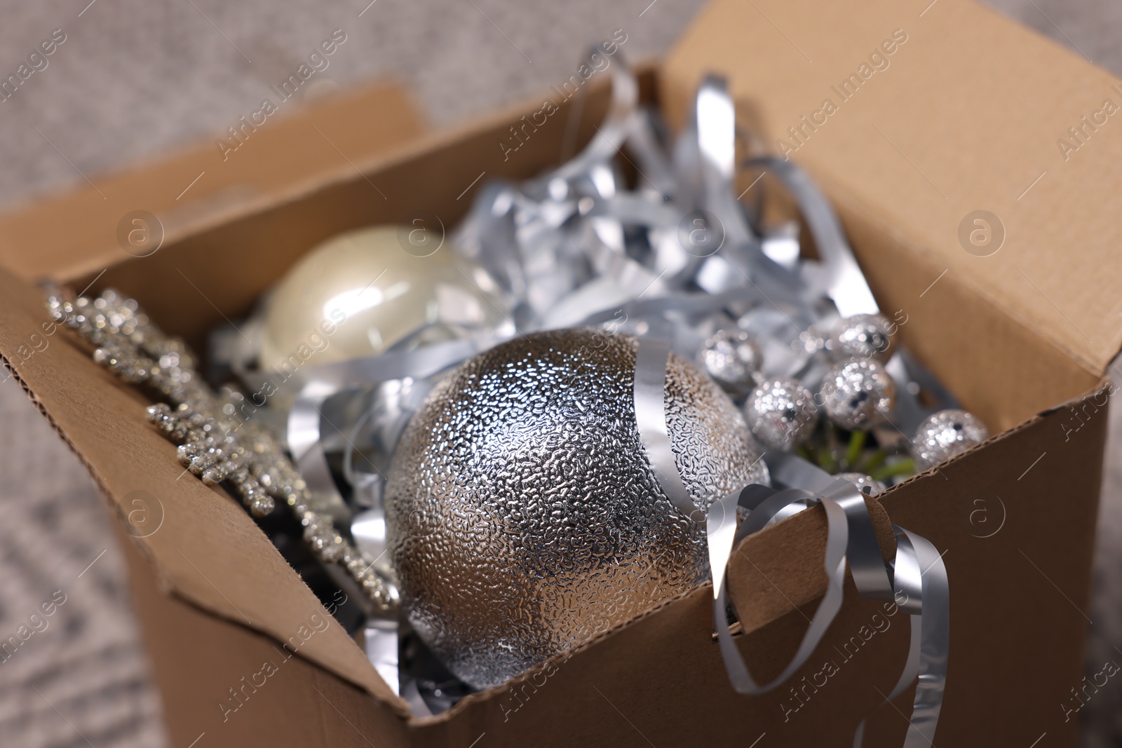 Photo of Shiny tinsels, baubles and other Christmas decor in cardboard box against blurred background, closeup