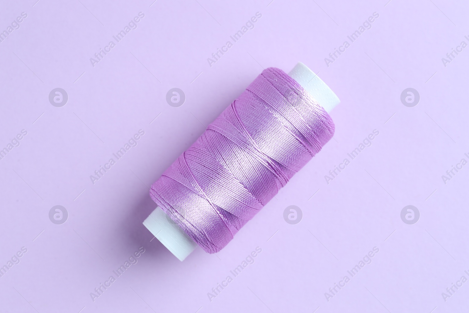 Photo of Spool of violet sewing thread on lilac background, top view