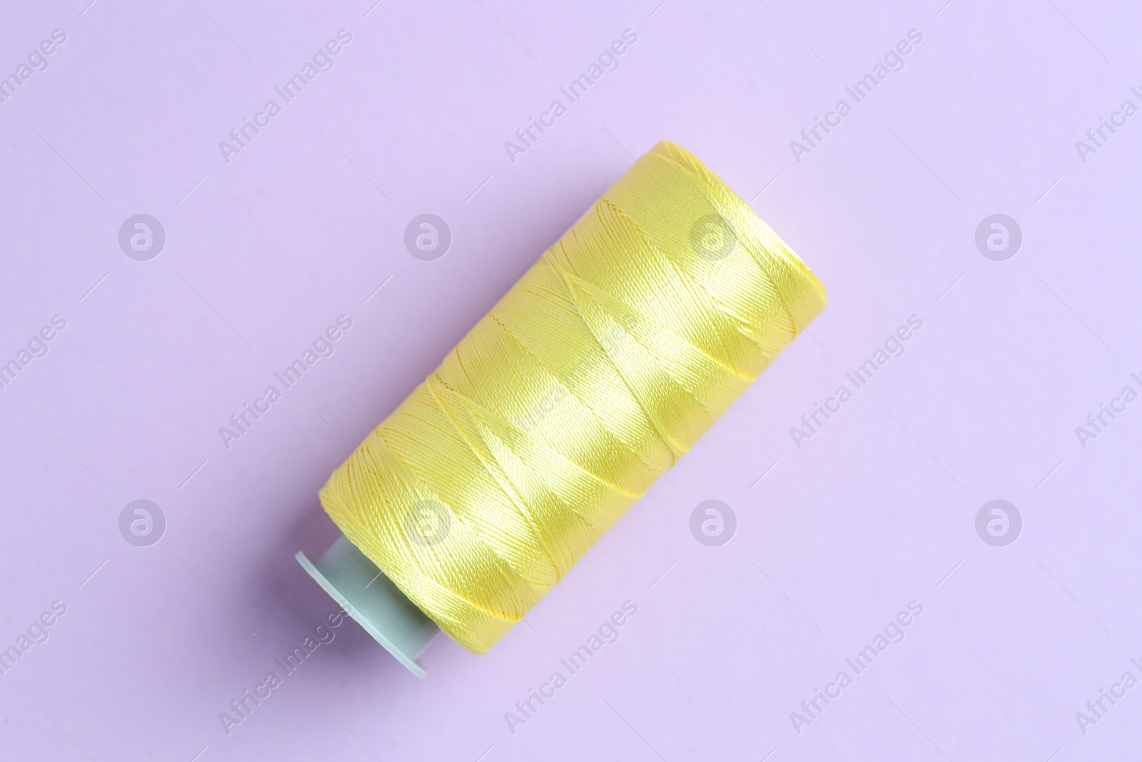 Photo of Spool of yellow sewing thread on lilac background, top view