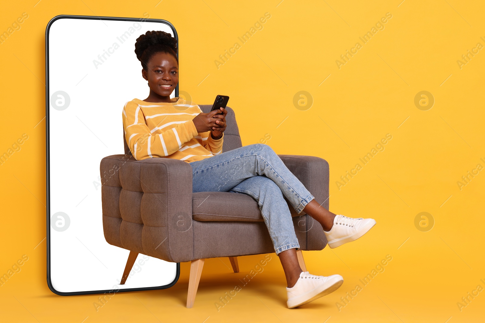 Image of Happy woman with smartphone sitting in armchair on orange background. Big mobile phone with blank screen behind her