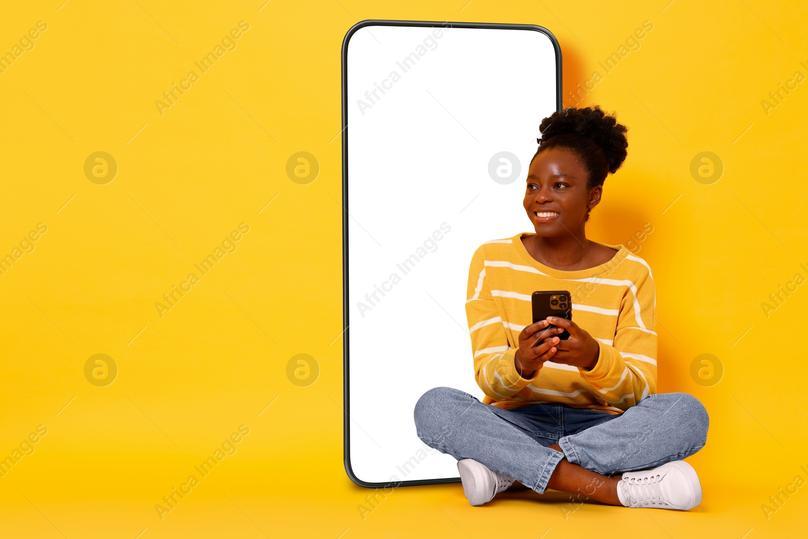 Image of Happy woman with smartphone sitting on orange background. Big mobile phone with blank screen behind her