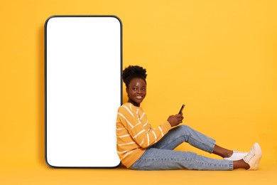 Image of Happy woman with smartphone leaning on big mobile phone with blank screen on orange background