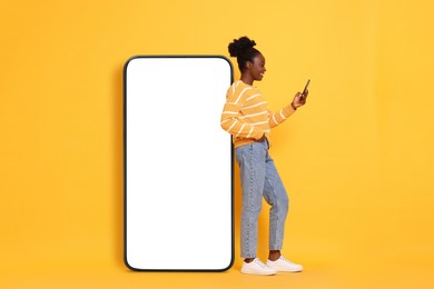 Image of Happy woman with smartphone leaning on big mobile phone with blank screen on orange background