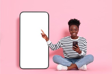 Image of Happy woman with smartphone pointing at big mobile phone with blank screen on pink background