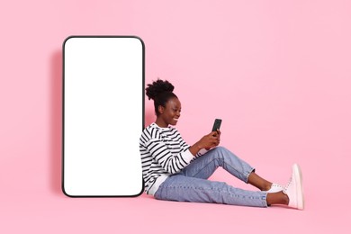 Image of Happy woman with smartphone leaning on big mobile phone with blank screen on pink background