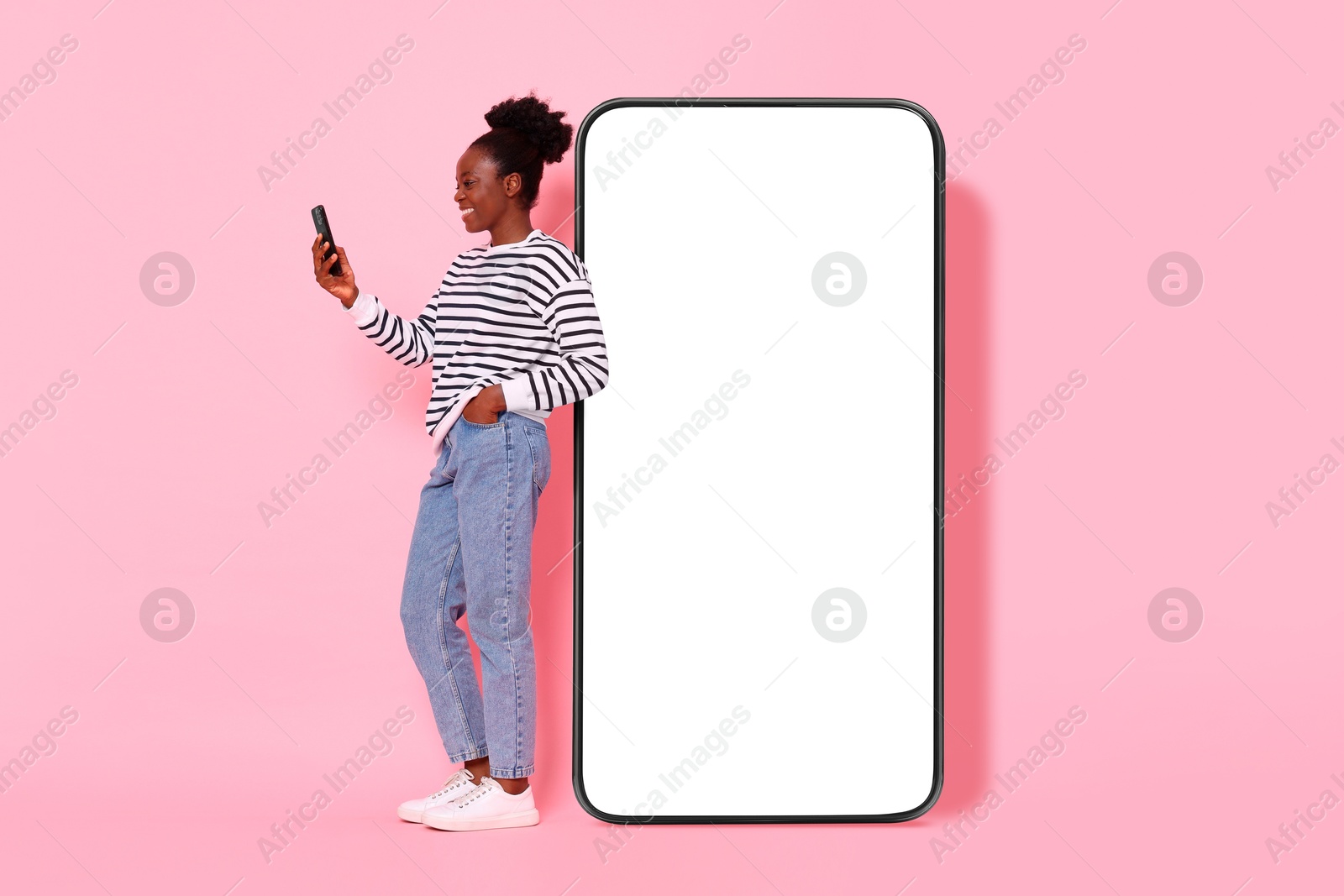 Image of Happy woman with smartphone leaning on big mobile phone with blank screen on pink background