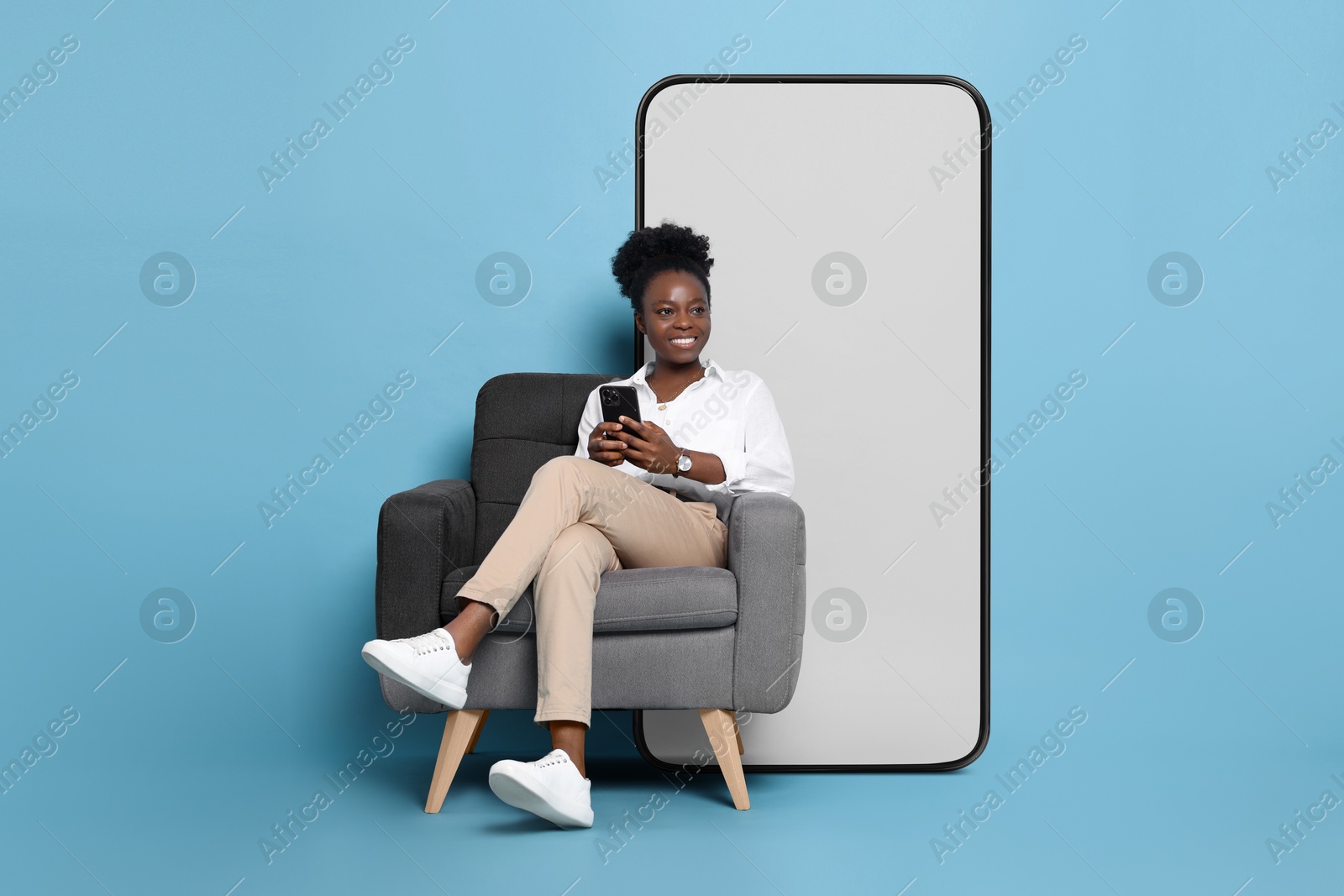 Image of Happy woman with smartphone sitting in armchair on light blue background. Big mobile phone with blank screen behind her