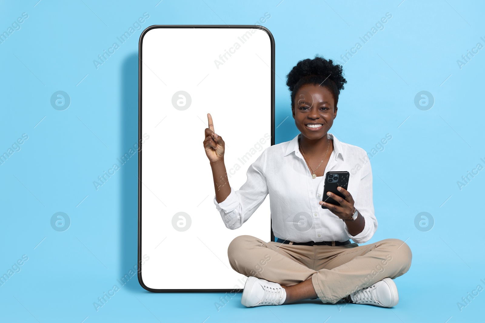 Image of Happy woman with smartphone pointing at big mobile phone with blank screen on light blue background