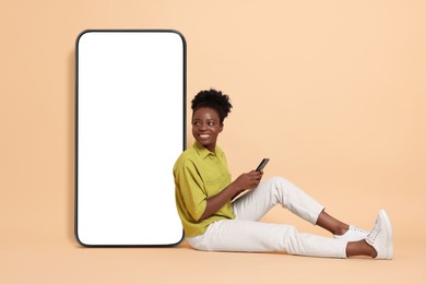 Image of Happy woman with smartphone leaning on big mobile phone with blank screen on beige background