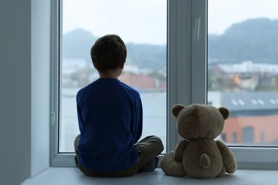 Photo of Autism concept. Lonely little boy with teddy bear near window at home, back view