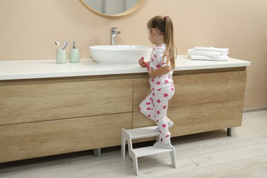 Photo of Little girl with toothbrush standing on step stool near bathroom vanity indoors