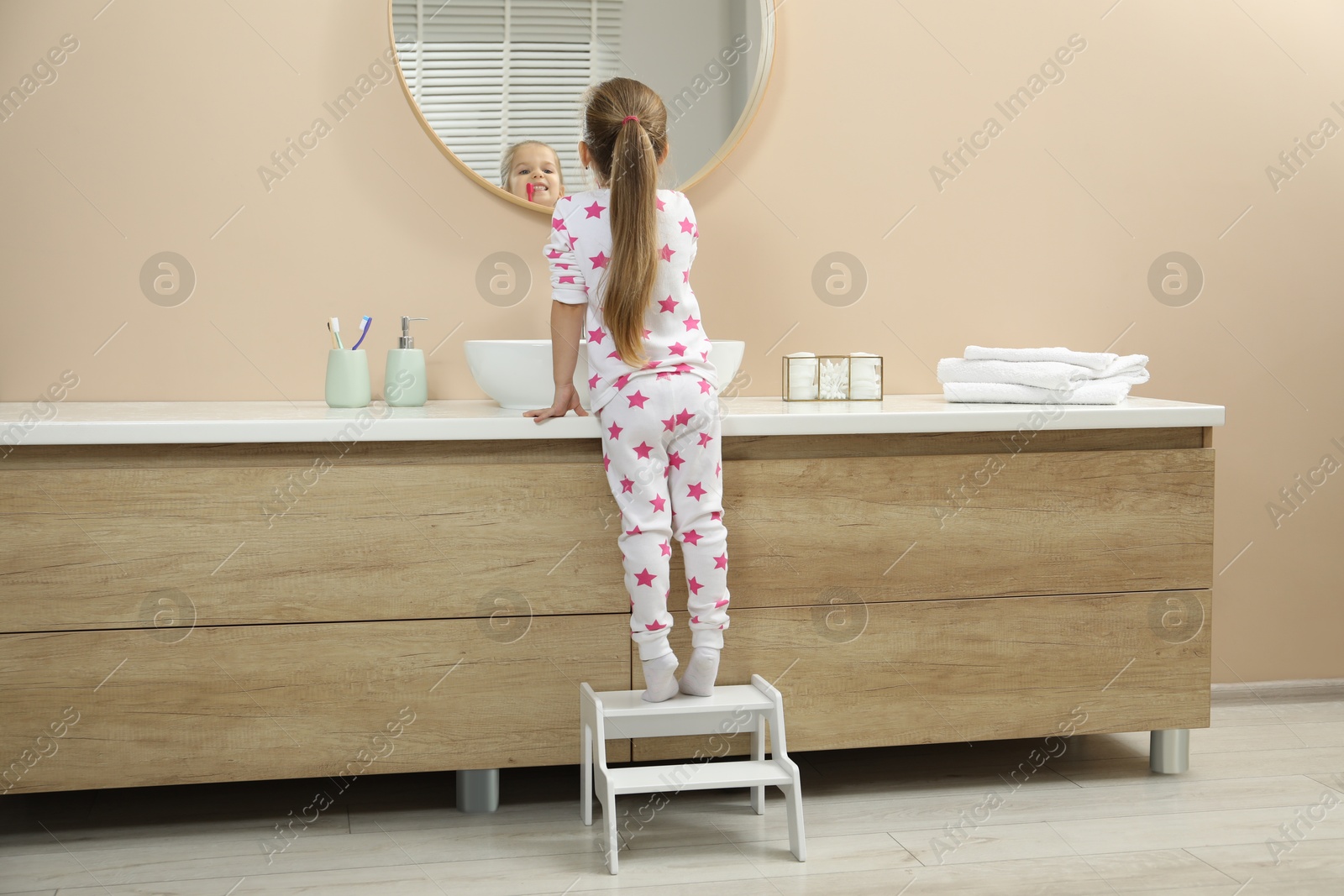 Photo of Little girl with toothbrush standing on step stool near bathroom vanity indoors, back view