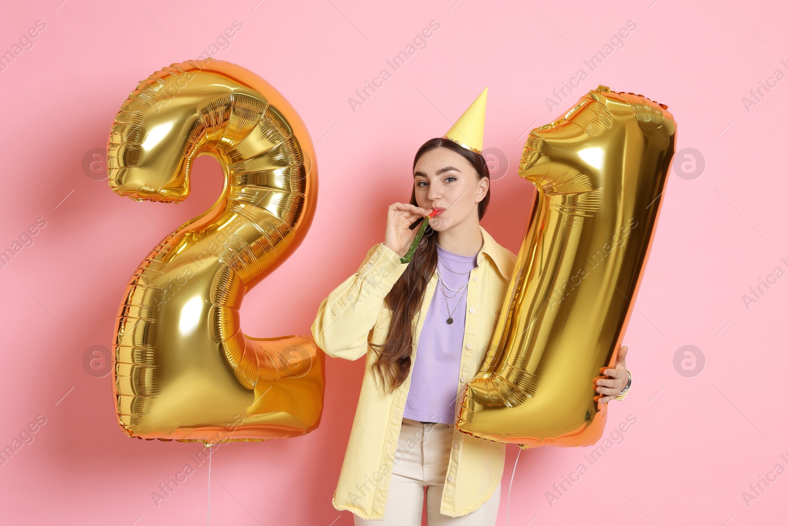 Photo of Coming of age party - 21st birthday. Happy young woman with number shaped balloons, hat and blower on pink background