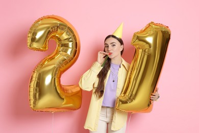 Photo of Coming of age party - 21st birthday. Happy young woman with number shaped balloons, hat and blower on pink background