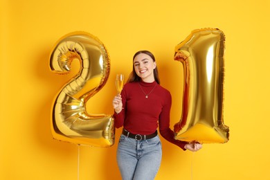 Photo of Coming of age party - 21st birthday. Happy young woman with number shaped balloons and glass of sparkling wine on yellow background