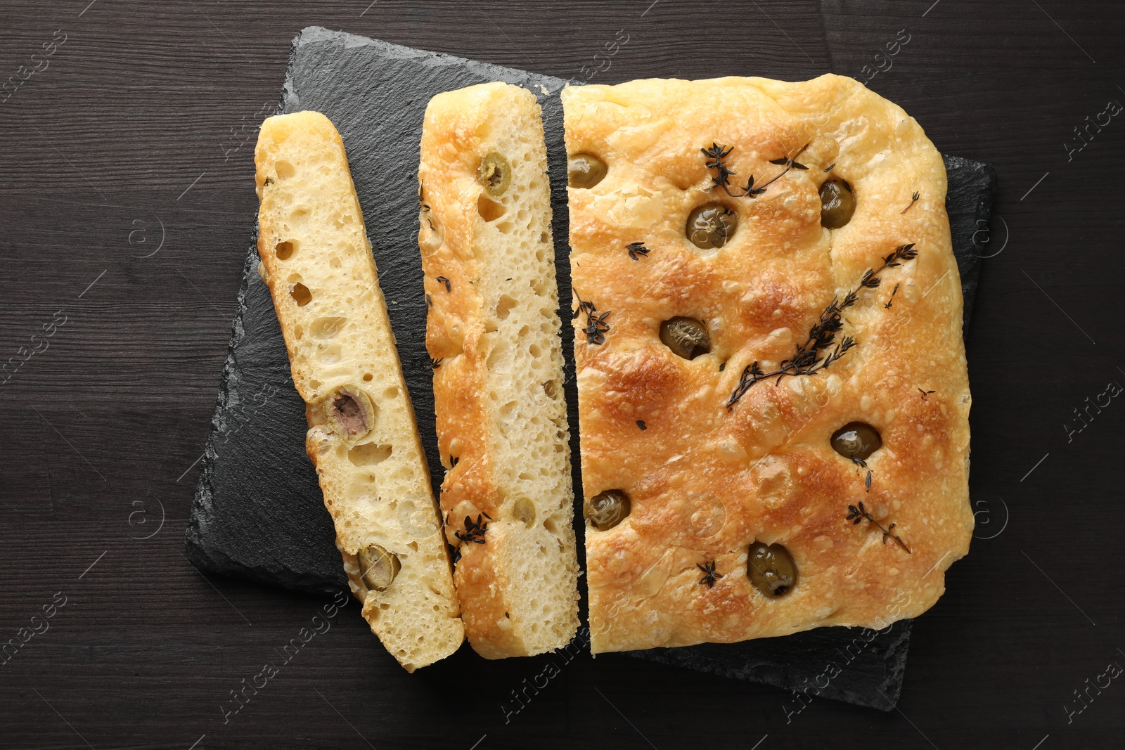 Photo of Pieces of delicious focaccia bread with olives and thyme on dark wooden table, top view