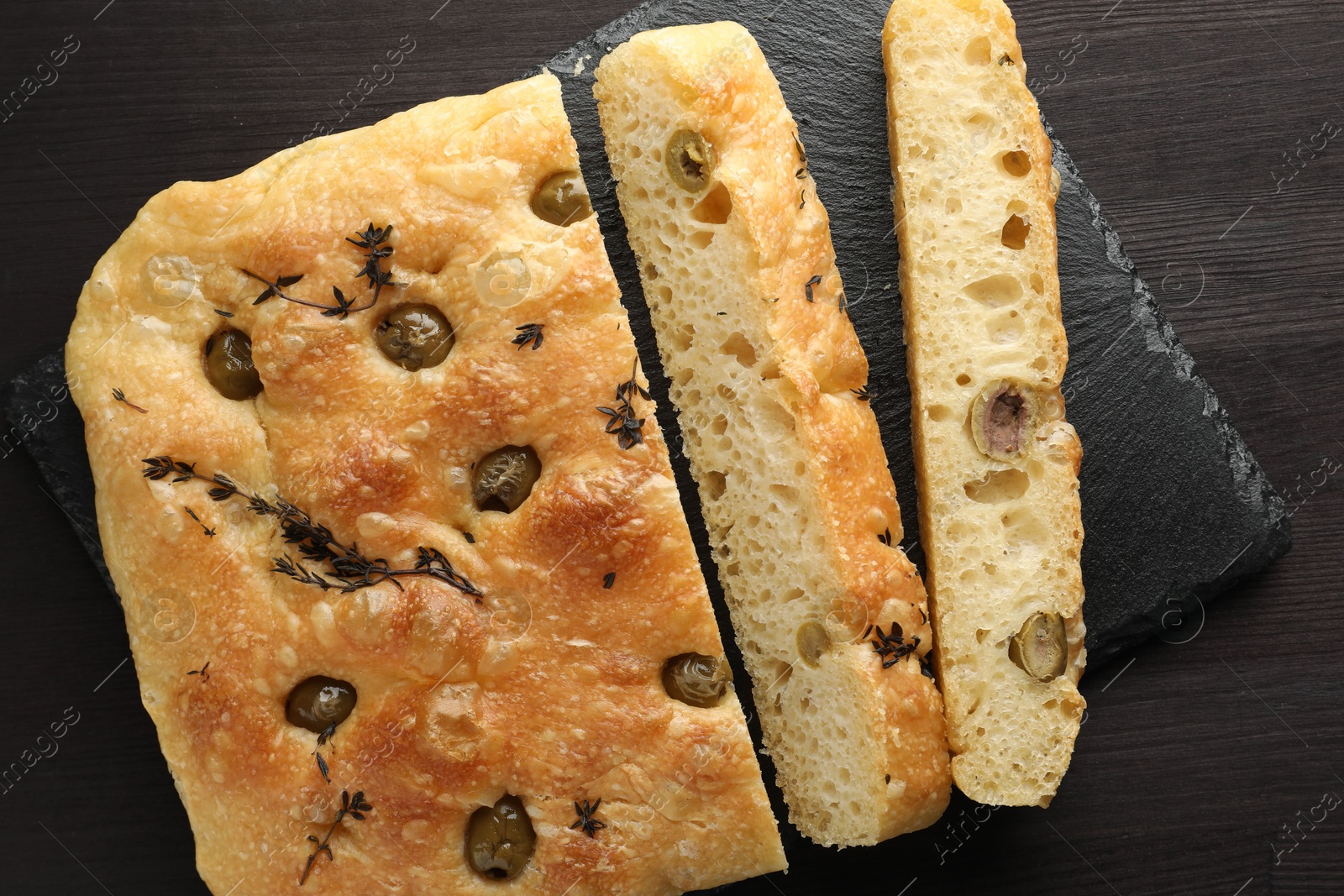 Photo of Pieces of delicious focaccia bread with olives and thyme on dark wooden table, top view