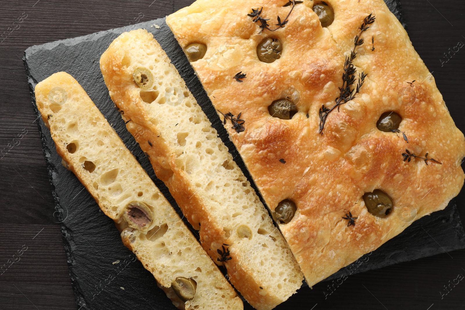 Photo of Pieces of delicious focaccia bread with olives and thyme on dark wooden table, top view