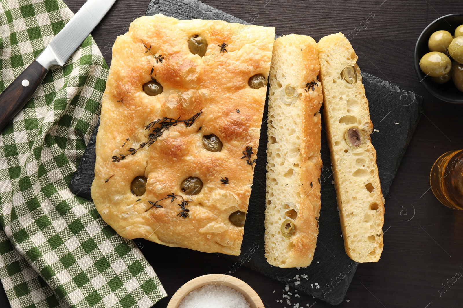 Photo of Pieces of delicious focaccia bread with olives, thyme, salt and knife on dark wooden table, flat lay