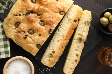 Photo of Pieces of delicious focaccia bread with olives, thyme, salt and knife on dark wooden table, flat lay