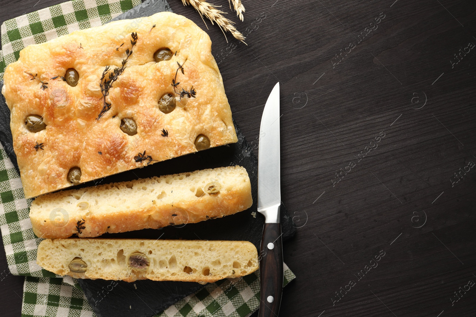 Photo of Pieces of delicious focaccia bread with olives, thyme, spikes and knife on dark wooden table, flat lay. Space for text