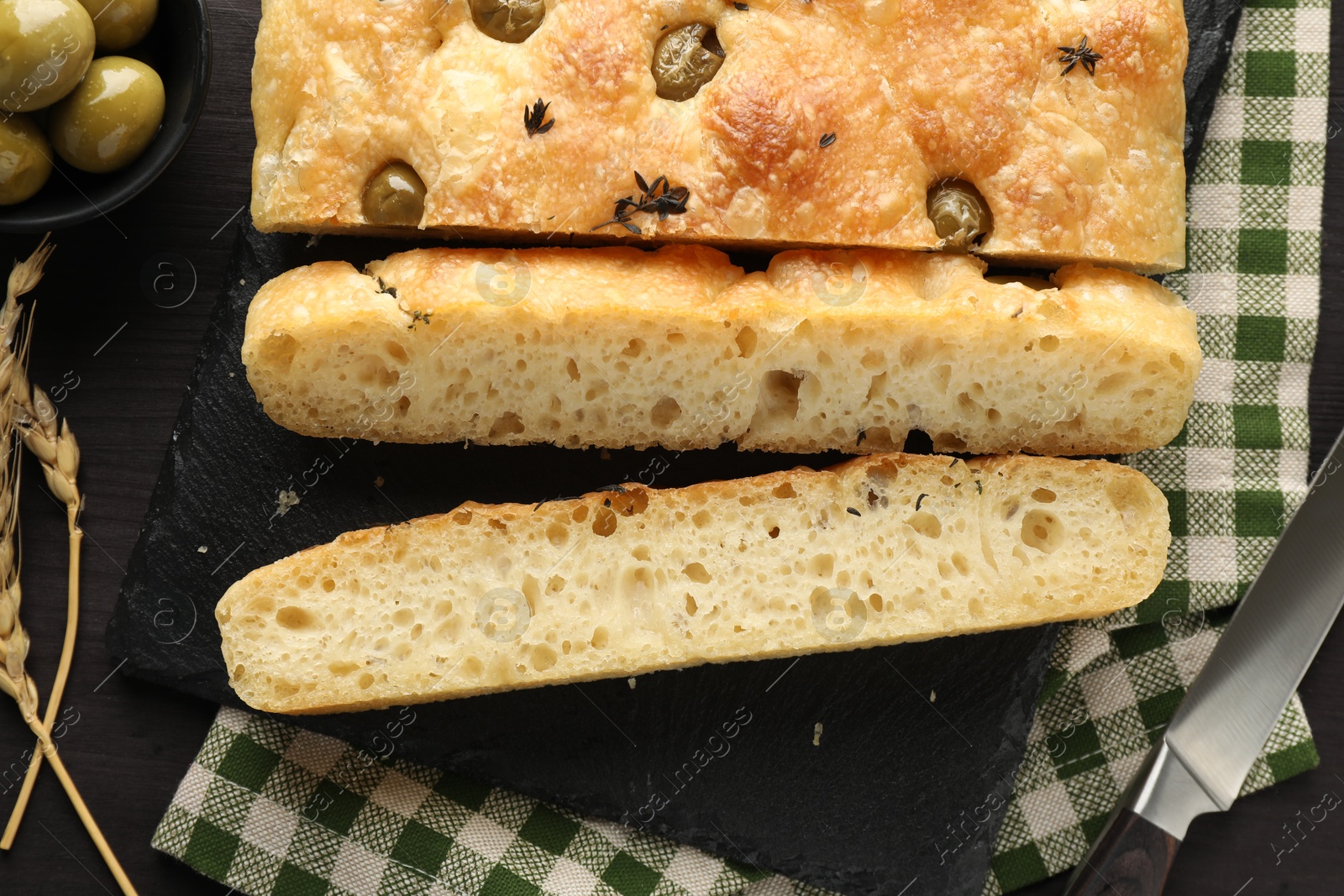 Photo of Pieces of delicious focaccia bread with olives, thyme, spikes and knife on dark wooden table, flat lay