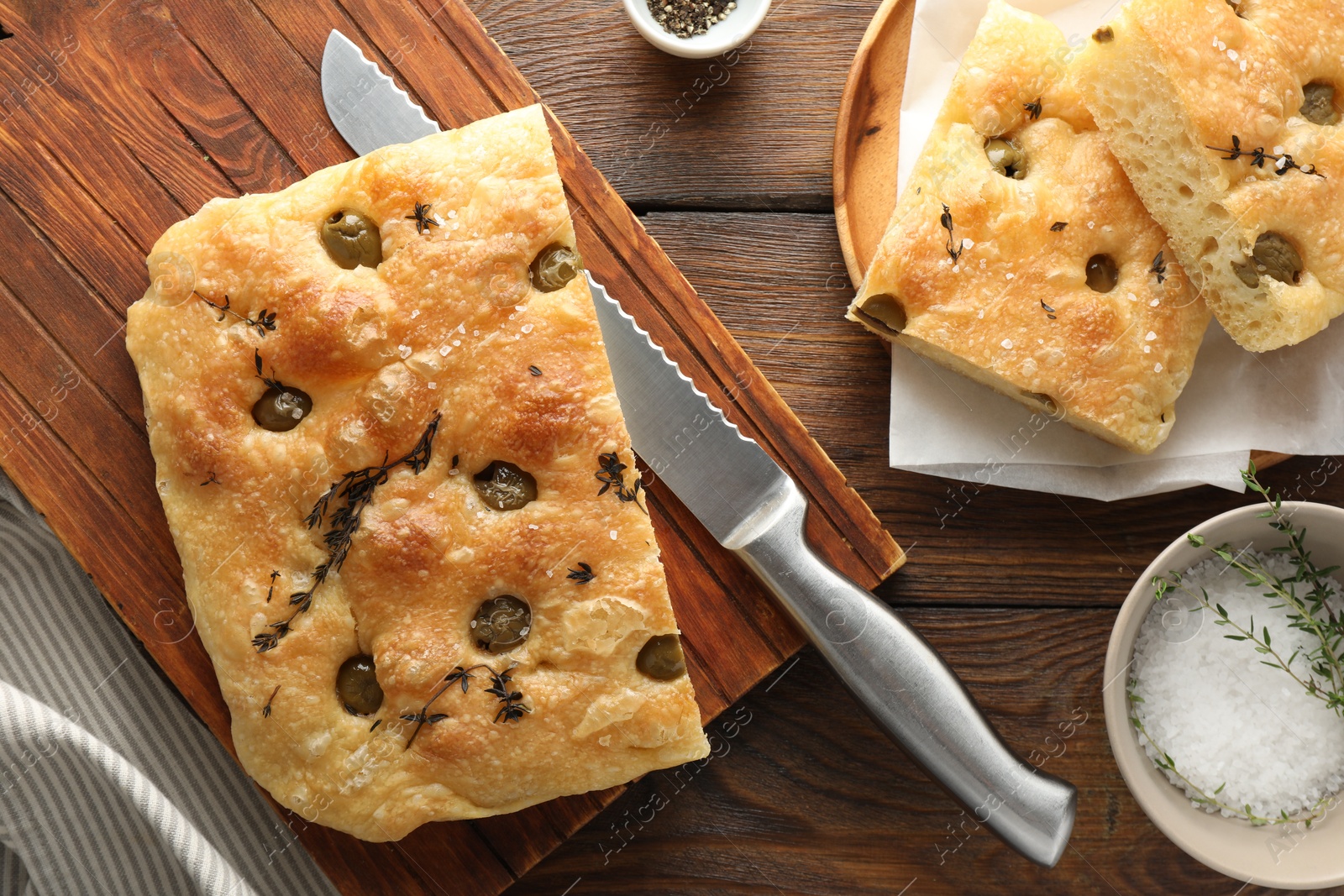 Photo of Pieces of delicious focaccia bread with olives, thyme, salt and knife on wooden table, flat lay