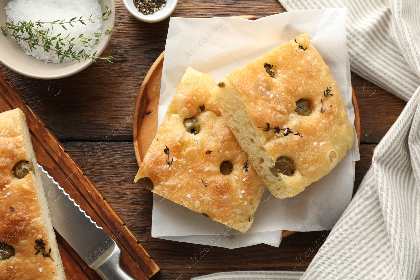 Photo of Pieces of delicious focaccia bread with olives, thyme, salt and knife on wooden table, flat lay