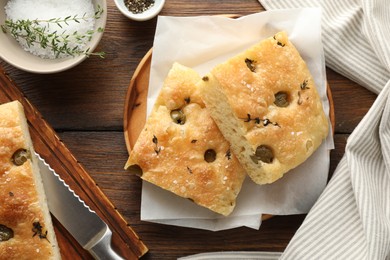 Photo of Pieces of delicious focaccia bread with olives, thyme, salt and knife on wooden table, flat lay