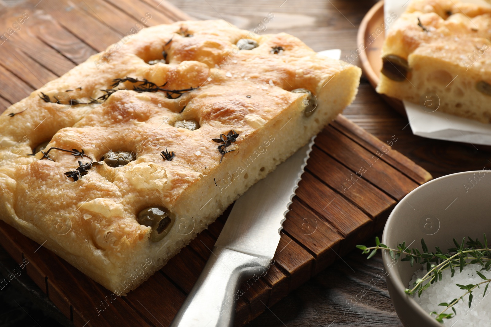 Photo of Pieces of delicious focaccia bread with olives, thyme, salt and knife on wooden table, closeup