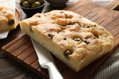 Photo of Piece of delicious focaccia bread with olives, thyme, salt and knife on wooden table, closeup