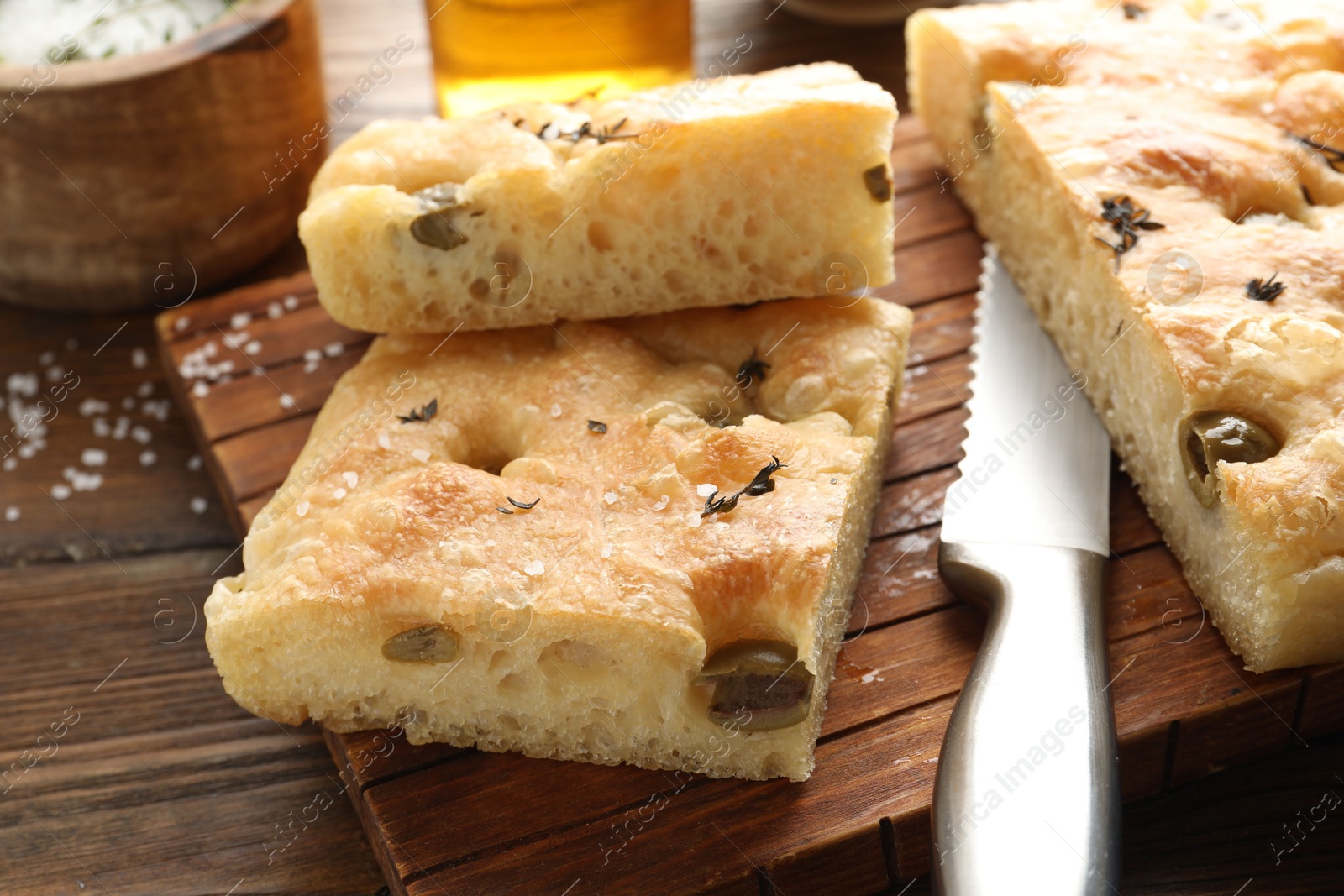 Photo of Pieces of delicious focaccia bread with olives, thyme, salt and knife on wooden table, closeup