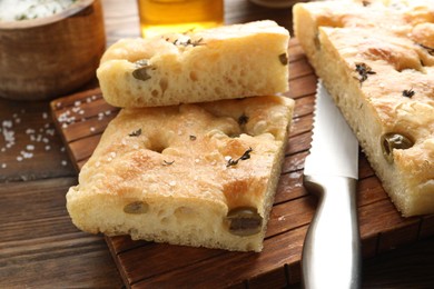 Photo of Pieces of delicious focaccia bread with olives, thyme, salt and knife on wooden table, closeup