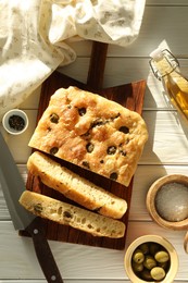 Photo of Pieces of delicious focaccia bread with olives, thyme, spices and knife on white wooden table, flat lay