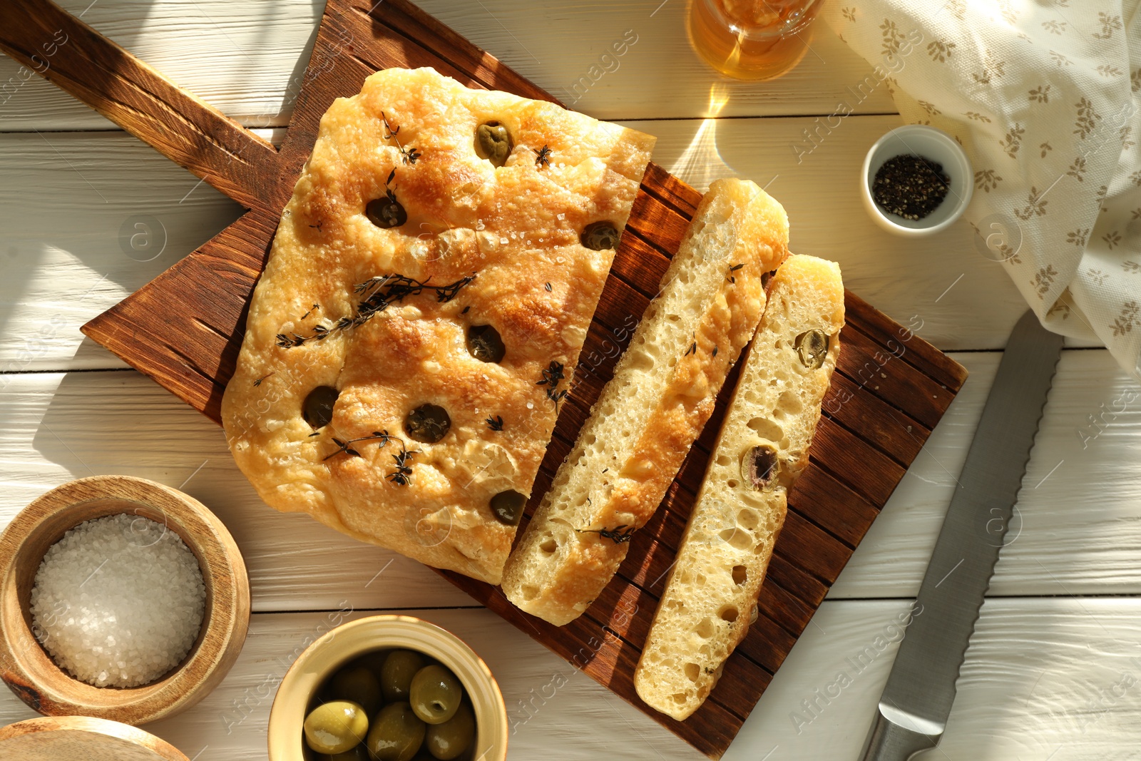 Photo of Pieces of delicious focaccia bread with olives, thyme, spices and knife on white wooden table, flat lay