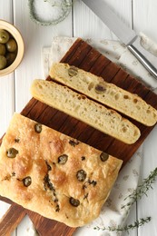 Photo of Pieces of delicious focaccia bread with olives, thyme, salt and knife on white wooden table, flat lay