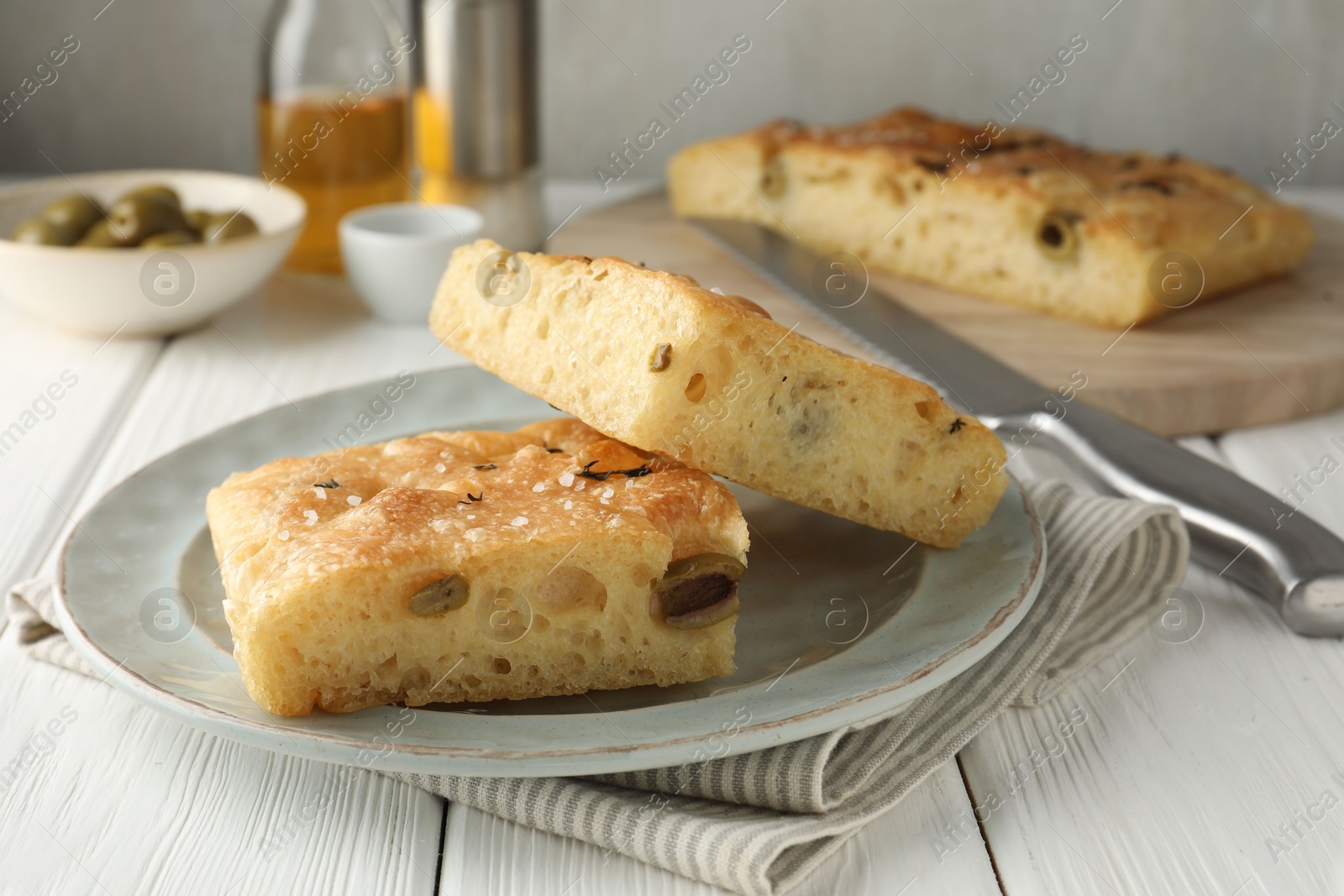 Photo of Pieces of delicious focaccia bread with olives, thyme, salt and knife on white wooden table, closeup