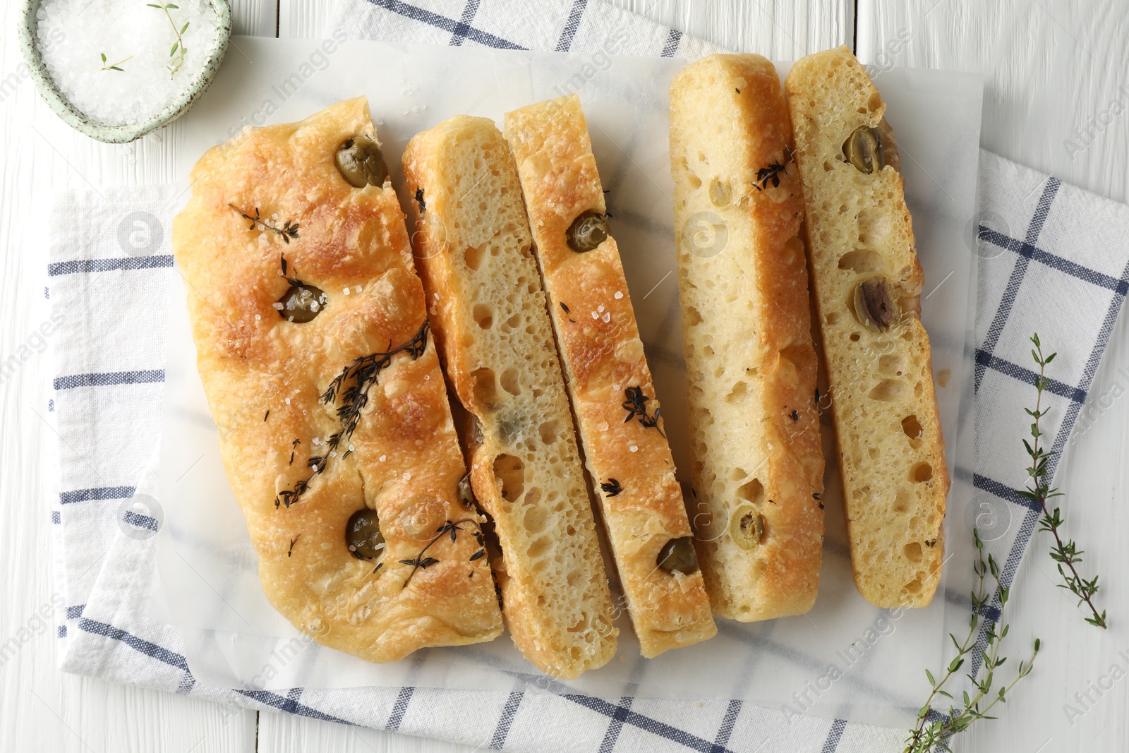 Photo of Slices of delicious focaccia bread with olives, thyme and salt on white wooden table, flat lay