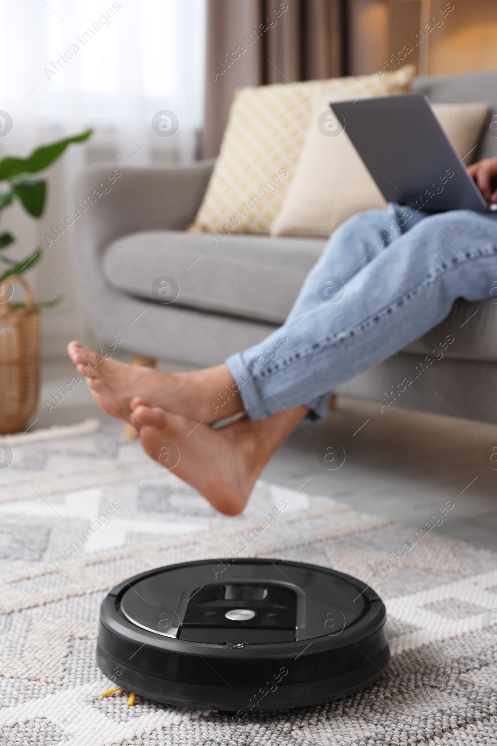 Photo of Young man using laptop while robotic vacuum cleaner vacuuming floor at home, closeup