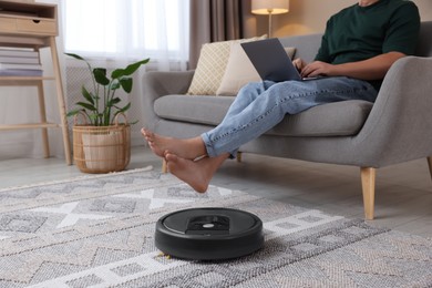 Photo of Young man using laptop while robotic vacuum cleaner vacuuming floor at home, closeup