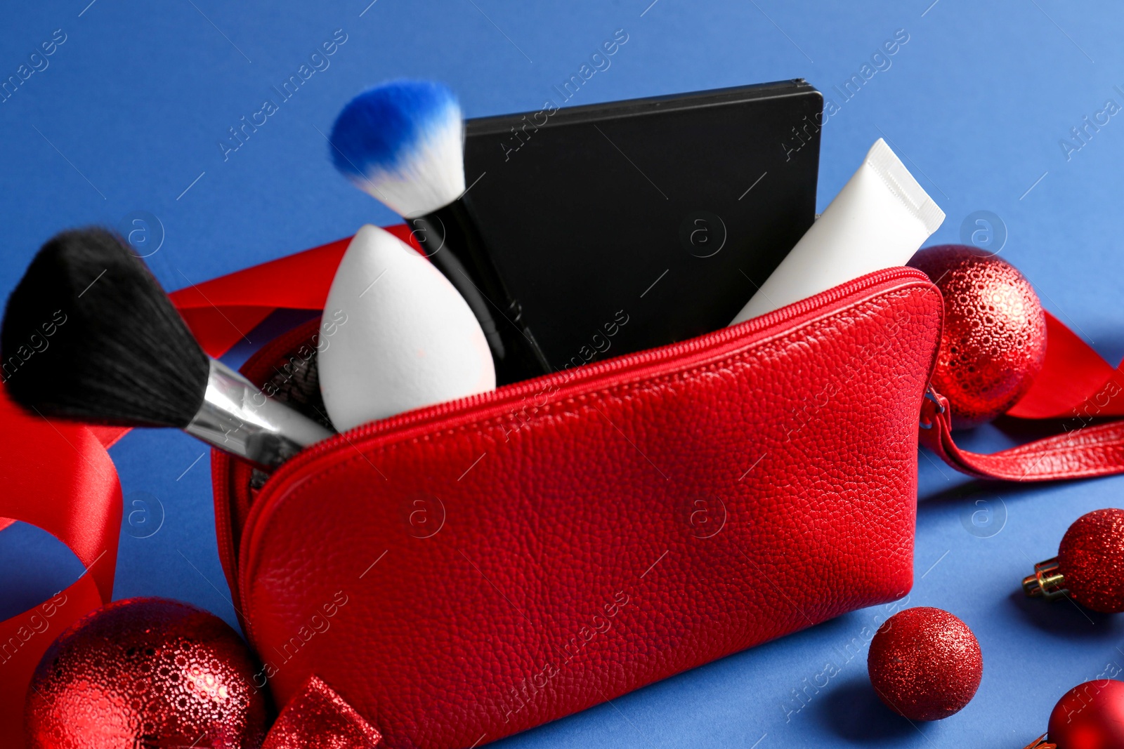 Photo of Christmas gift. Decorative cosmetics in red makeup bag and festive decor on blue background, closeup