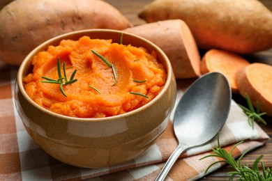 Photo of Tasty mashed sweet potato with rosemary in bowl, fresh vegetables and spoon on table, closeup