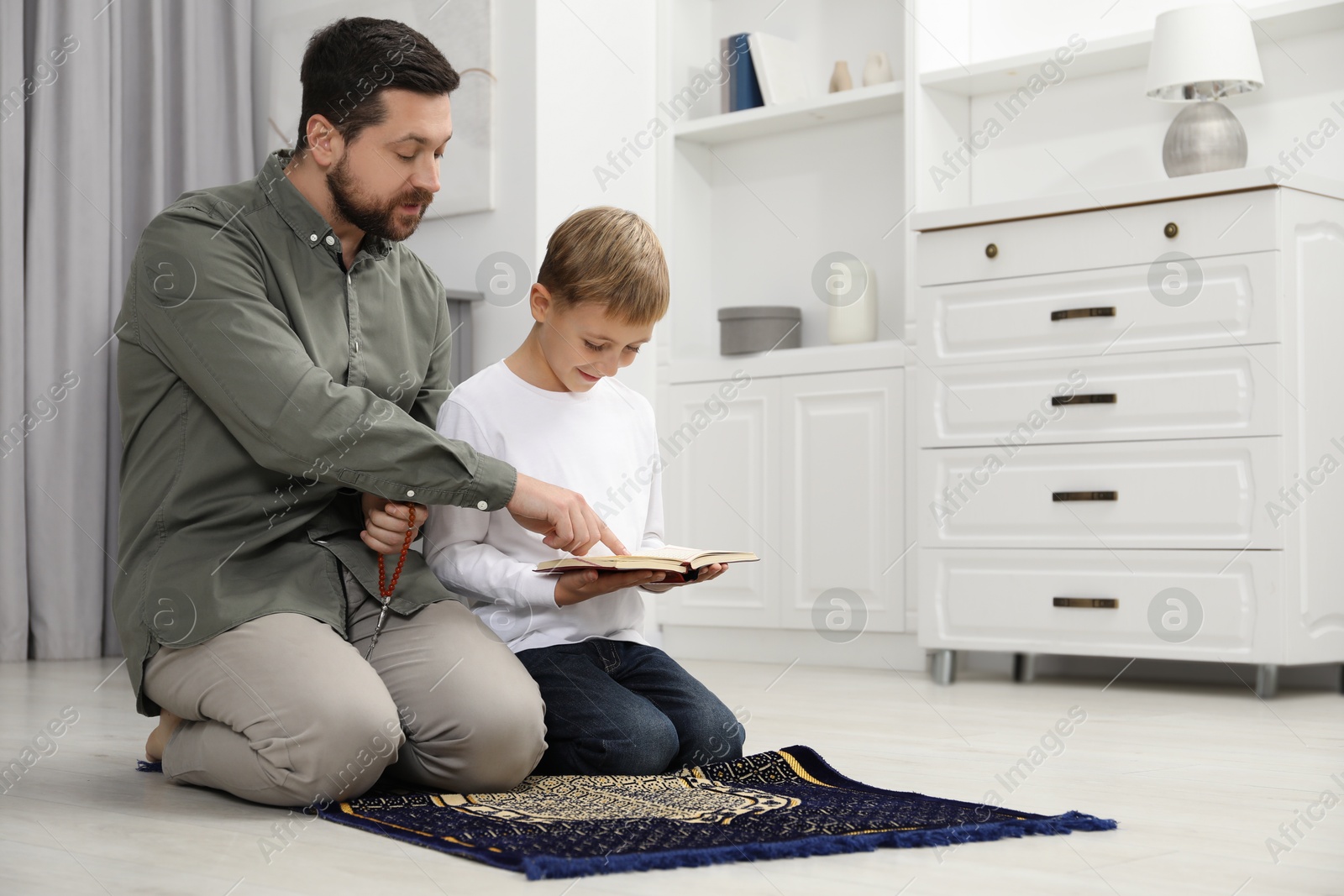 Photo of Muslim man with his son reading Quran and praying on mat at home, space for text