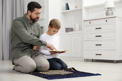 Photo of Muslim man with his son reading Quran and praying on mat at home, space for text