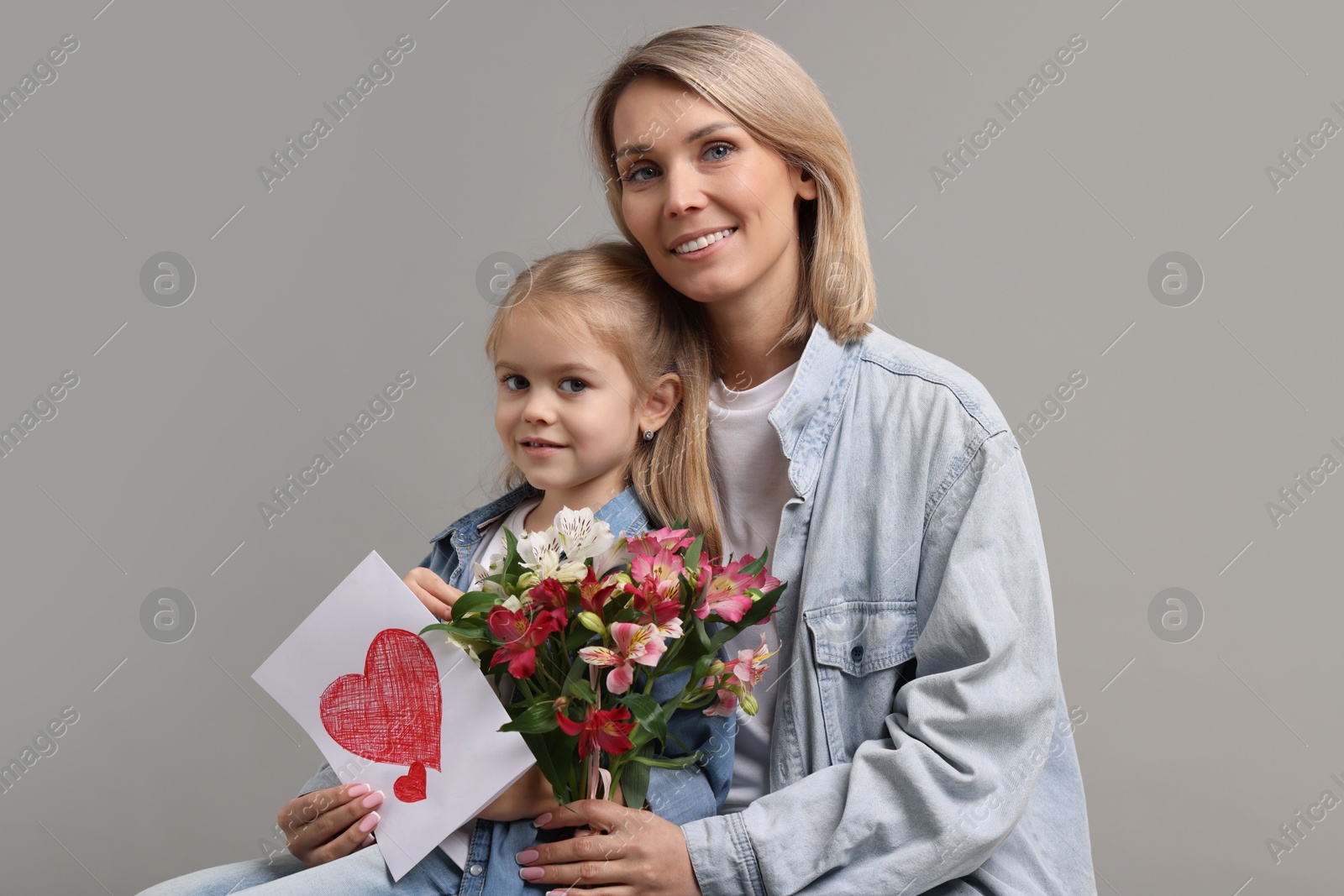 Photo of Happy woman with her daughter, bouquet of alstroemeria flowers and greeting card on gray background. Mother's Day celebration